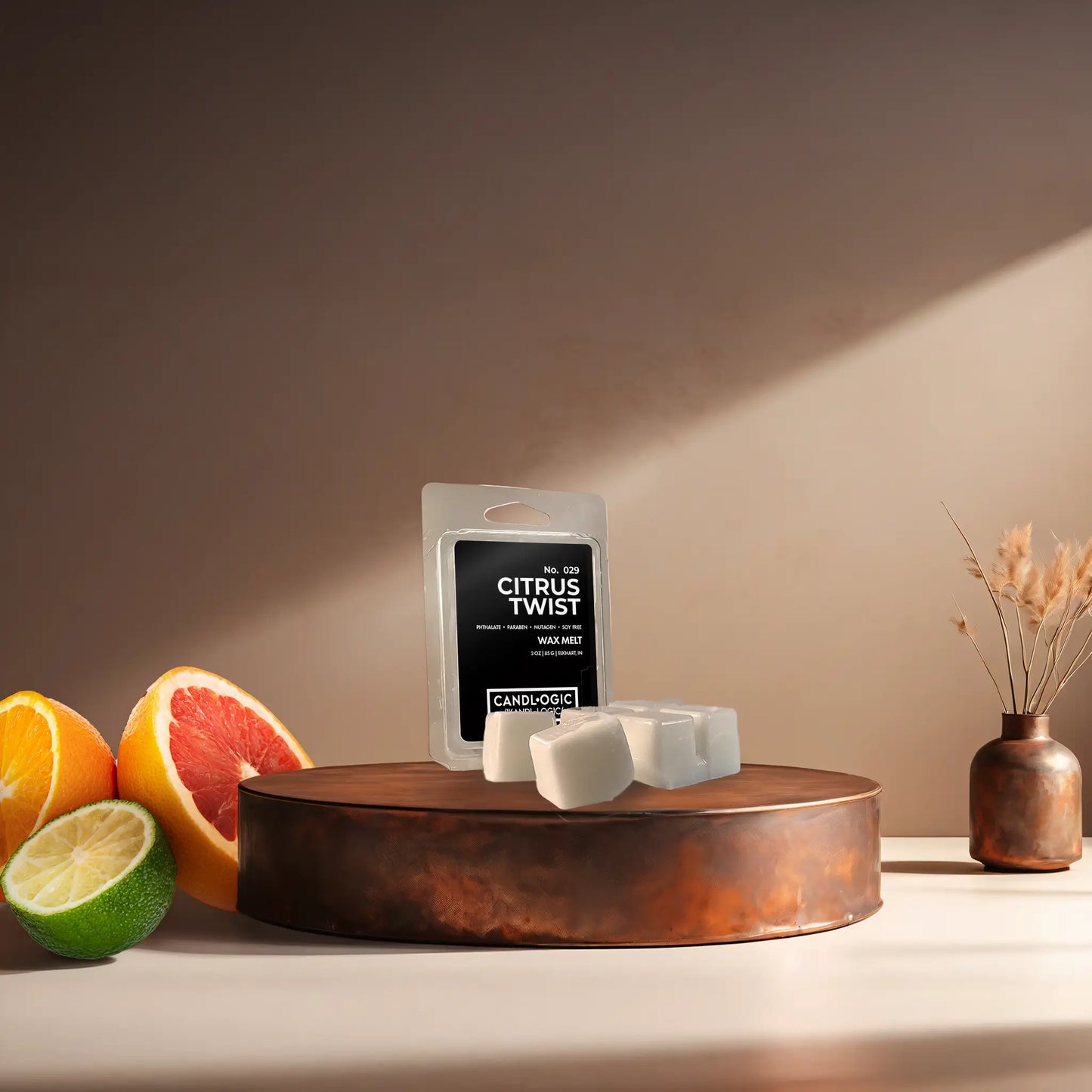 Candl•ogic’s wax melt cubes with packaging labeled ‘No. 029 Citrus Twist’ on a copper tray with citrus and a copper vase in the background.
