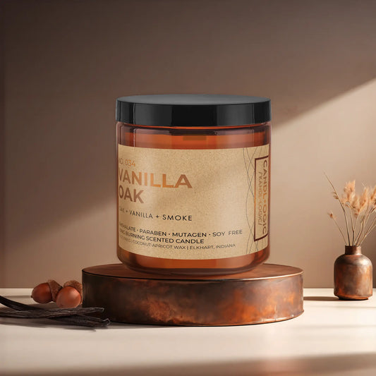 Candl•ogic’s scented candle with a wood wick in an amber glass 16 oz. jar labeled ‘No. 034 Vanilla Oak’ on a copper tray with acorns and vanilla beans and a copper vase in the background.