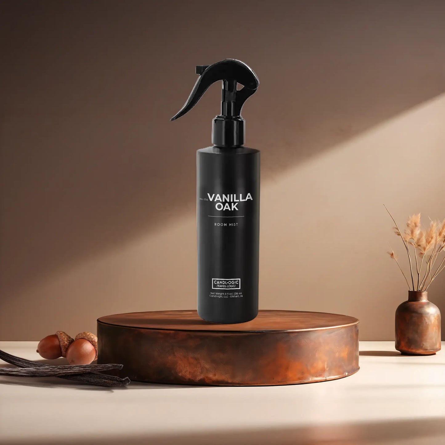 Candl•ogic’s 8 oz. room mist in a black spray bottle labeled ‘No. 034 Vanilla Oak’ on a copper tray with acorns and vanilla beans and a copper vase in the background.