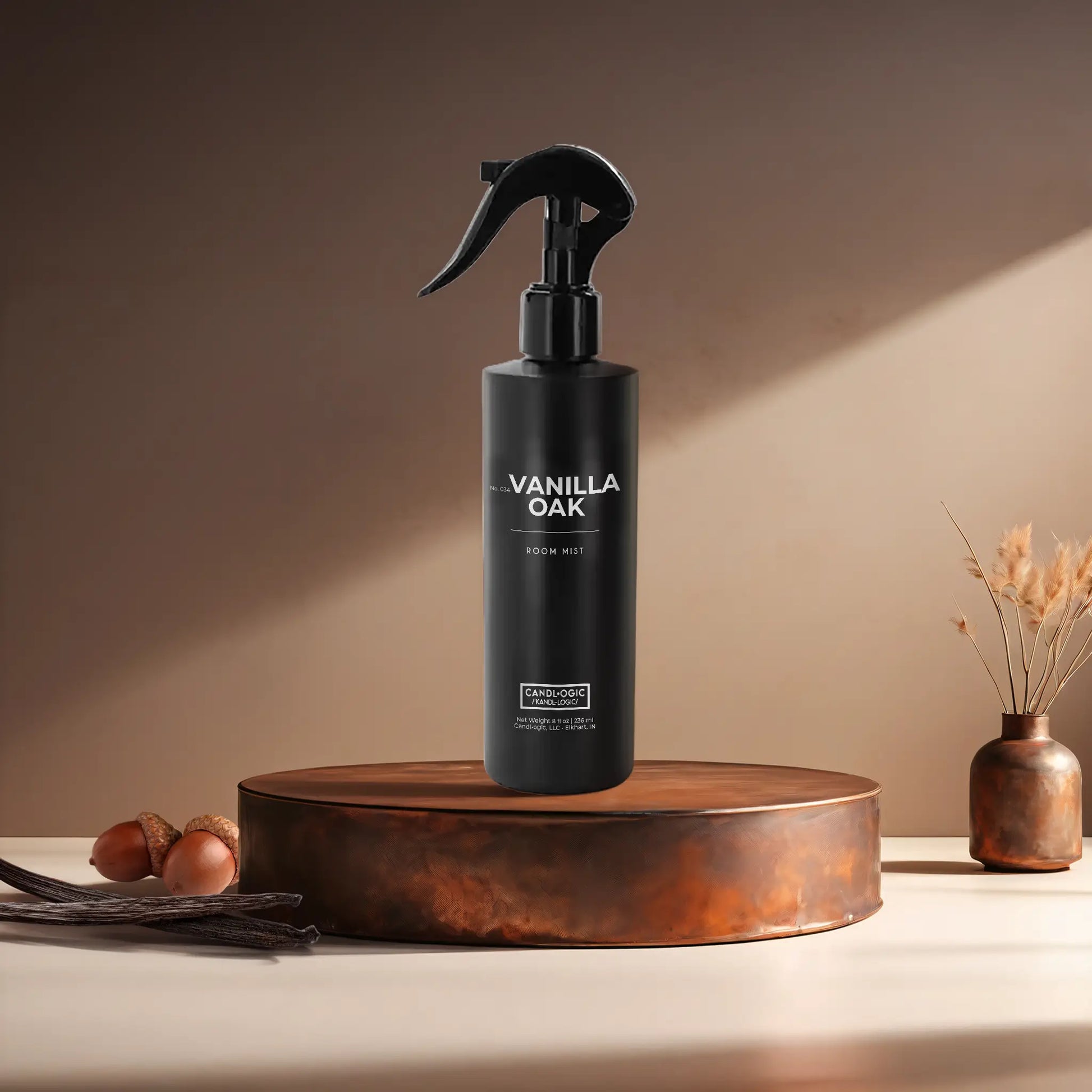 Candl•ogic’s 8 oz. room mist in a black spray bottle labeled ‘No. 034 Vanilla Oak’ on a copper tray with acorns and vanilla beans and a copper vase in the background.