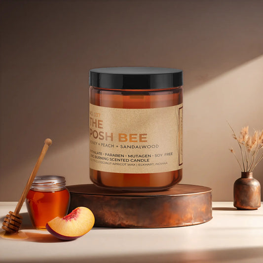 Candl•ogic's scented candle with a wood wick in an amber glass 9 oz. jar labeled 'No. 037 The Posh Bee ' on a copper tray with a small honey pot with wooden honey dipper and slice of a peach with a copper flower vase in the background.