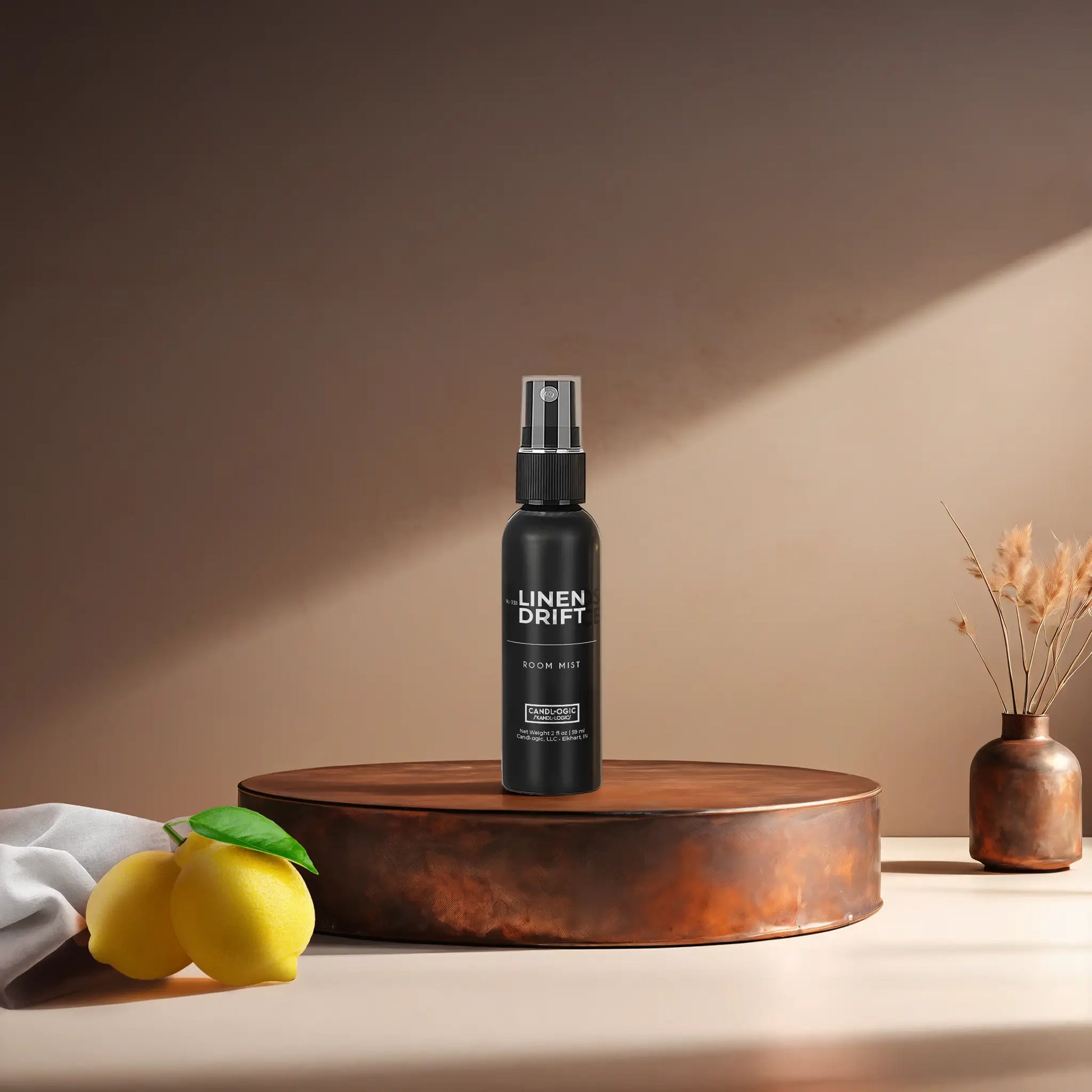 Candl•ogic's 2 oz. room mist in a black spray bottle labeled 'No. 038 Linen Drift' on a copper tray with a white fabric piece and lemons and a vase in the background.