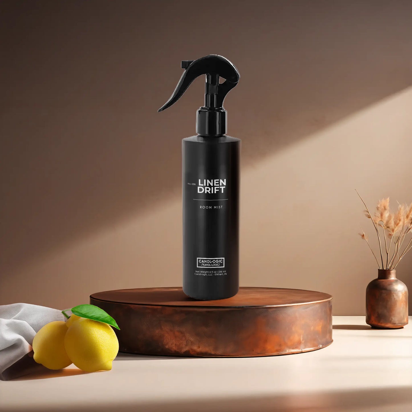 Candl•ogic's 8 oz. room mist in a black spray bottle labeled 'No. 038 Linen Drift' on a copper tray with a white fabric piece and lemons and a vase in the background.