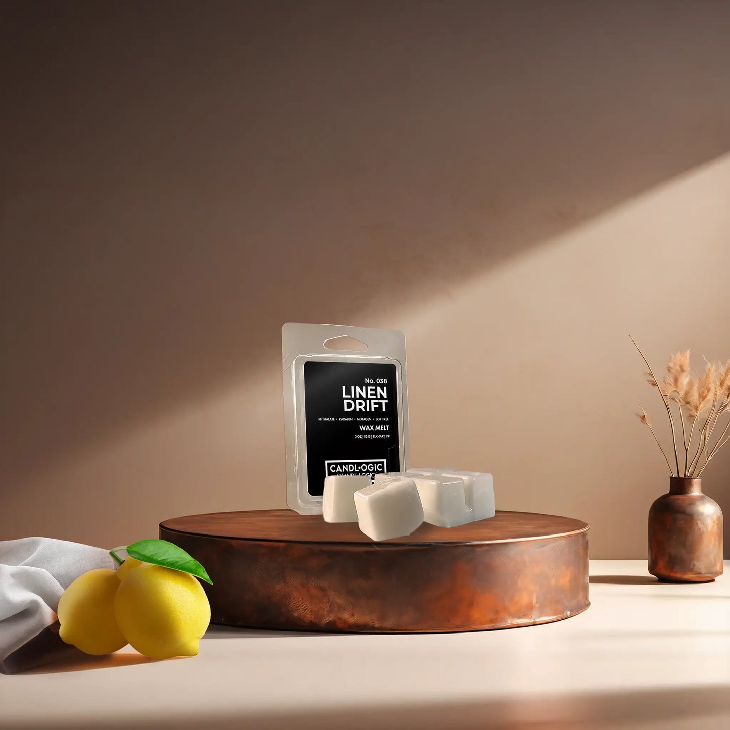 Candl•ogic's wax melt cubes with packaging labeled 'No. 038 Linen Drift' on a copper tray with a white fabric piece and lemons and a vase in the background.