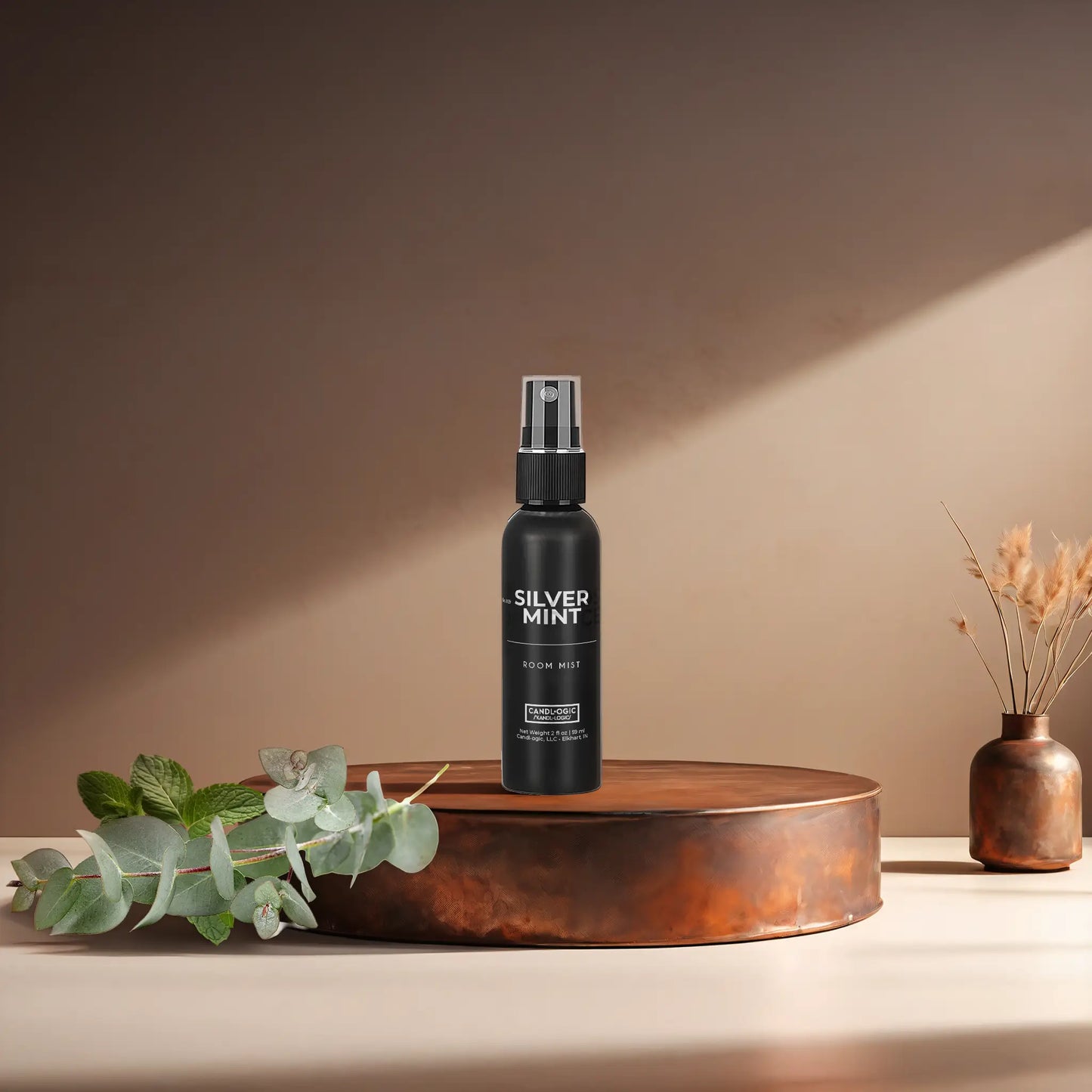 Candl•ogic's 2 oz. room mist in a black spray bottle labeled 'No. 039 Silver Mint' on a copper tray with eucalyptus and mint leaves and a vase in the background.