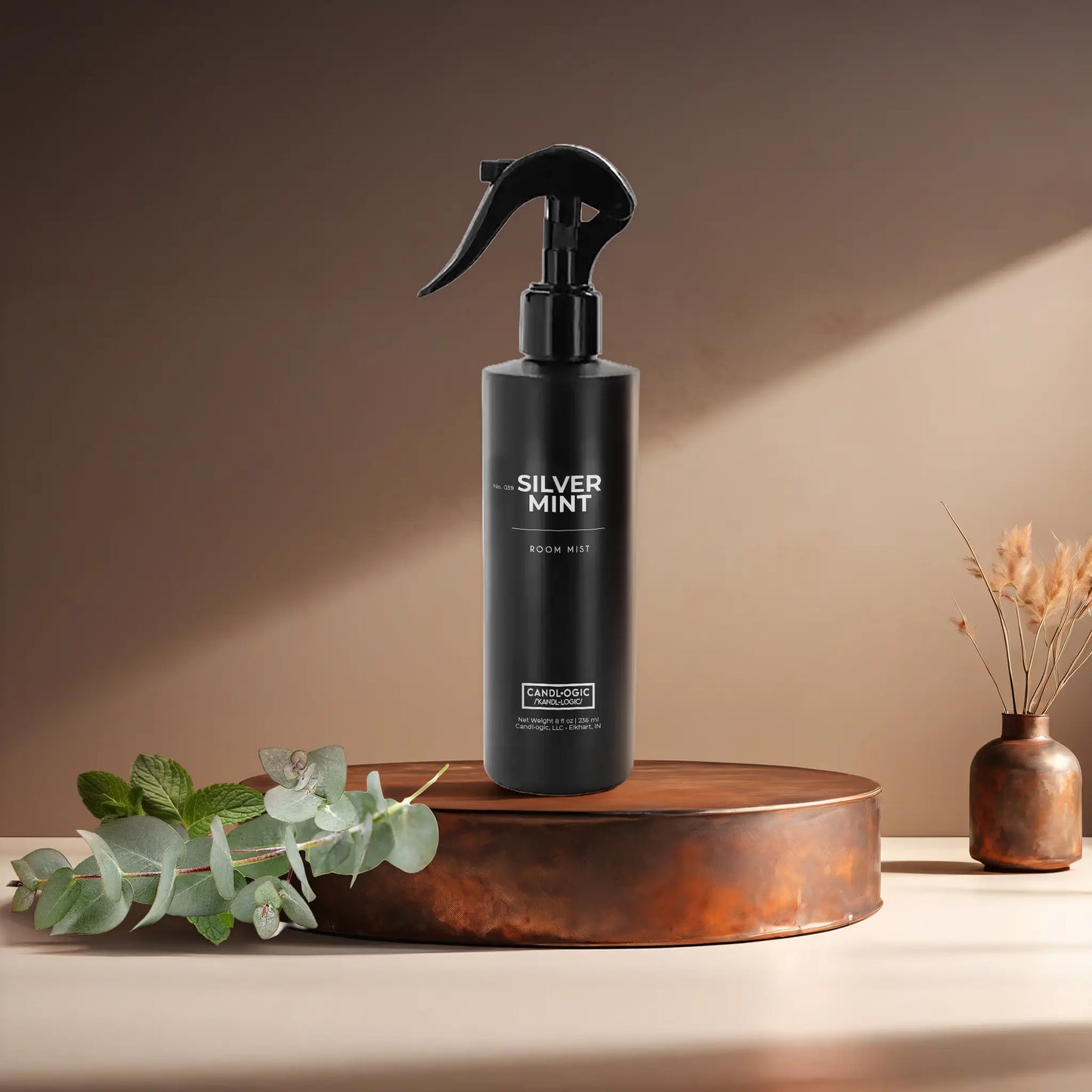Candl•ogic's 8 oz room mist in a black bottle labeled 'No. 039 Silver Mint' on a copper tray with eucalyptus and mint leaves and a vase in the background.