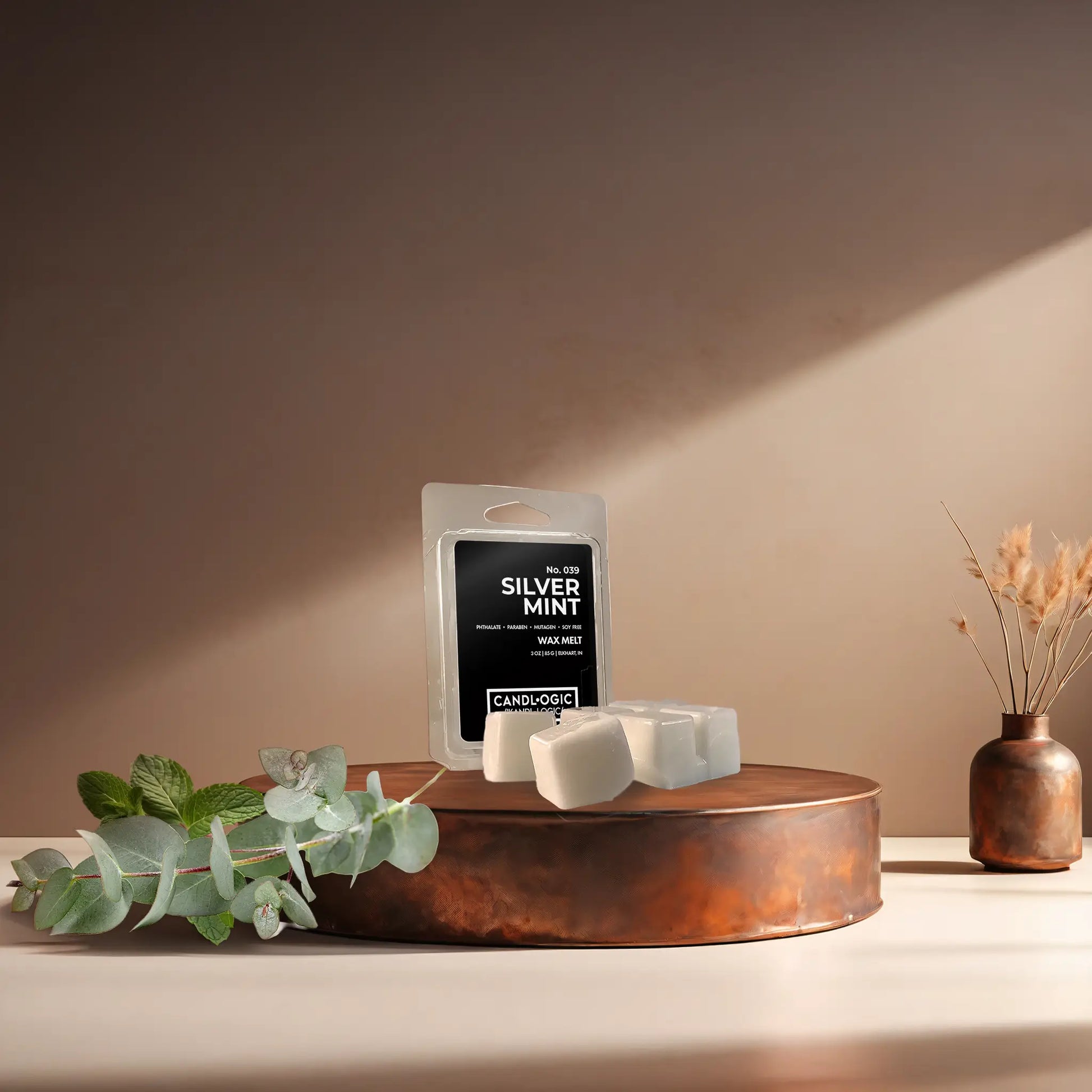 Candl•ogic's wax melt cubes and packaging labeled 'No. 039 Silver Mint' on a copper tray with eucalyptus and mint leaves and a vase in the background.