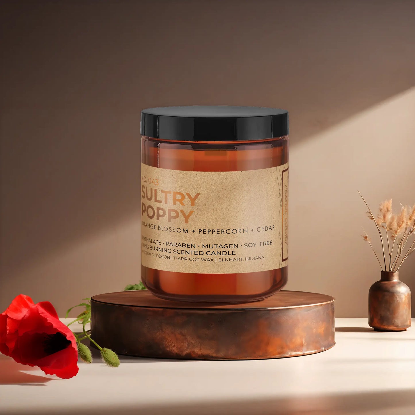 Candl•ogic's scented candle with a wood wick in an amber glass 9 oz. jar labeled 'No. 043 Sultry Poppy' on a copper tray with a red poppy flower and a vase in the background.