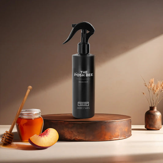 Candl•ogic's 8 oz. room mist in a black spray bottle labeled 'No. 037 The Posh Bee' on a copper tray with a small honey pot with wooden honey dipper and slice of a peach with a copper flower vase in the background.