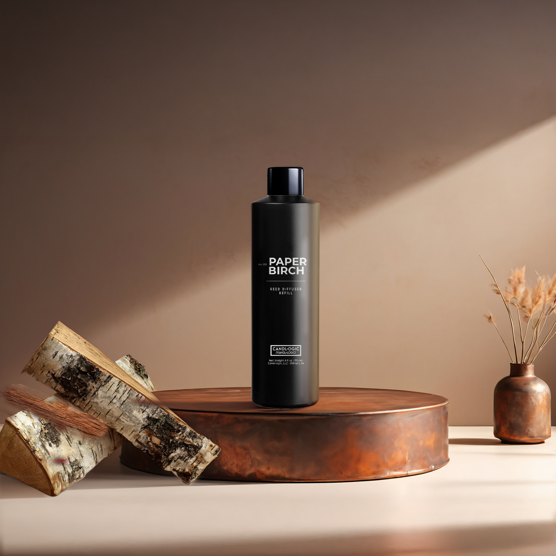 Candl•ogic’s 6 oz. reed diffuser refill solution in a black plastic bottle labeled ‘No. 012 Paper Birch’ on a copper tray with pieces of paper birch tree and a copper vase in the background.